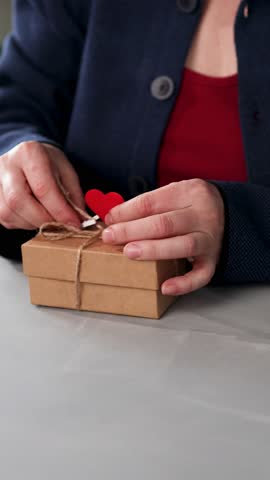 A person prepares a gift for someone special by tying a string around a box with a heart tag. This shows the effort put into making a heartfelt present for Valentine