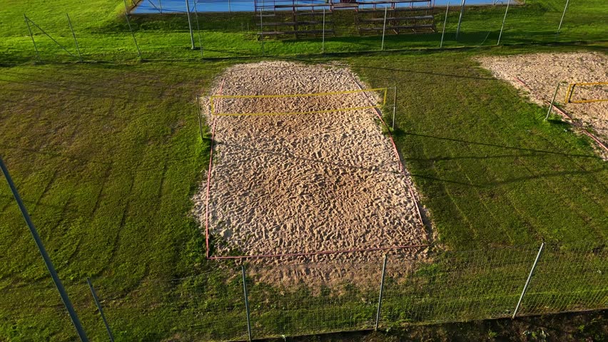 Drone view of an outdoor sand volleyball court surrounded by green fields, fences and evening shadows, showing textured sand, net structure and athletic space in warm natural sun light.