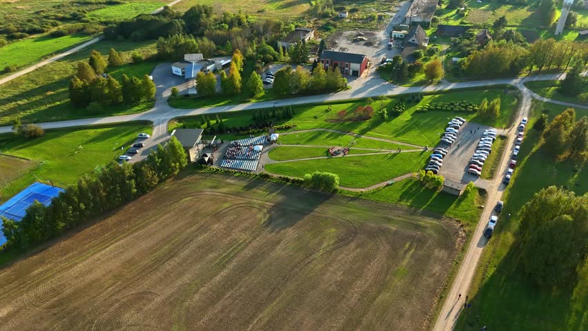 Aerial view of a rural outdoor park with a gathering by the stage, sports courts, green fields and full parking area on a sunny day. Scenic countryside layout lifestyle, travel and event.