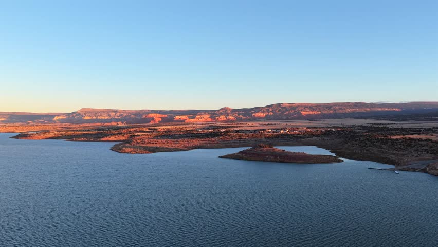 Stunning aerial view of a towering, sheer mesa rising dramatically from the blue water of Abiquiu Lake, New Mexico. The surrounding high desert landscape is scenic and evocative of the Southwest.