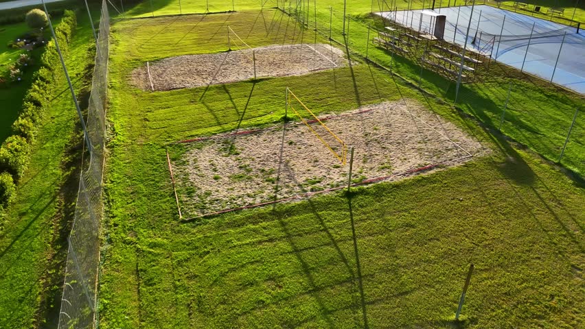 Aerial view of multiple sand volleyball courts surrounded by green fields, captured in warm evening light