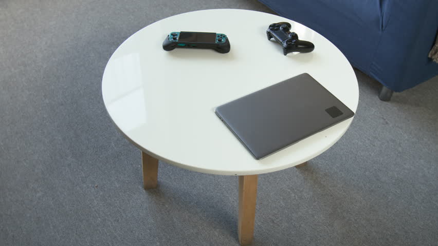 No people shot of video game controllers, digital tablet and laptop resting on coffee table and cozy couch with pillows in living room during day time, copy space