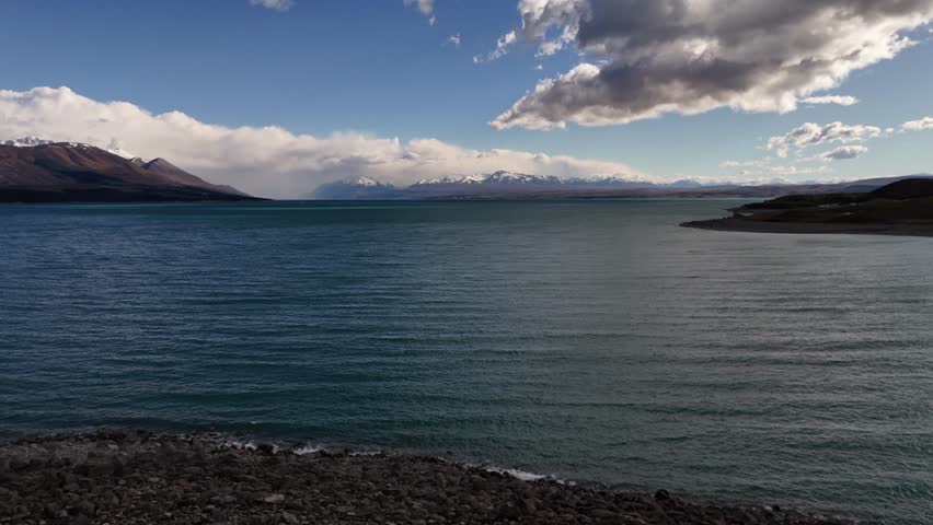 4K aerial drone view of the turquoise glacial water of Lake Pukaki in New Zealand. Rocky shoreline foreground with majestic Southern Alps mountains in the background under a cloudy sky