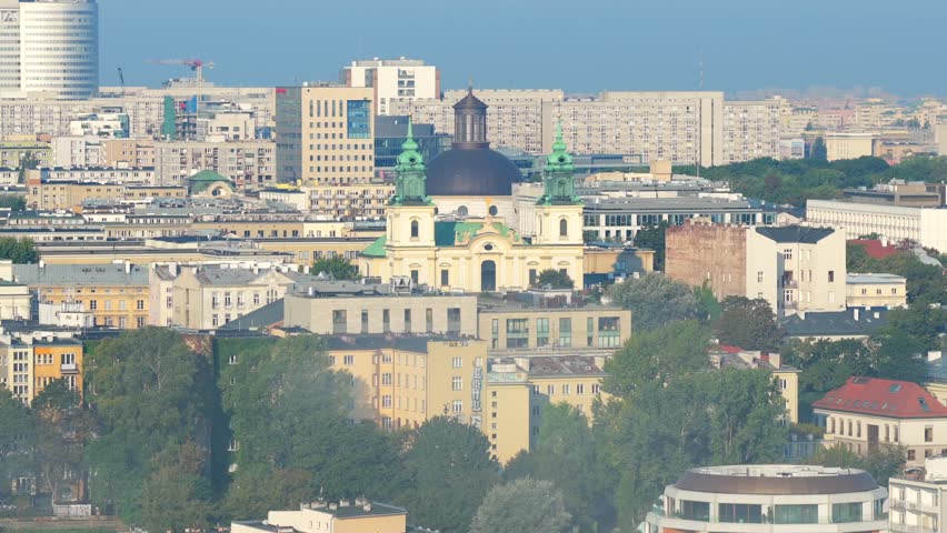 Aerial wide shot of Warsaw city with establishing shot of Holy Trinity Church as Church of Holy Cross tower rises in background. Dense city blocks and soft daylight with urban atmosphere in Poland.