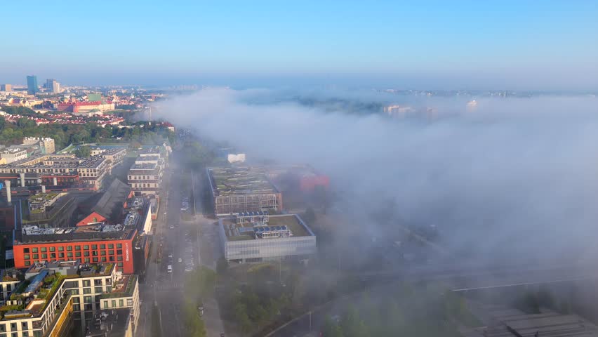 Aerial shot of Warsaw city as thick fog drifts across Vistula River and spreads into surrounding districts while bright morning light. city blocks emerging through soft mist. Wide shot