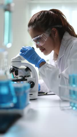Young female scientist wearing lab coat, safety glasses and gloves, carefully examining samples under a microscope in a modern laboratory.