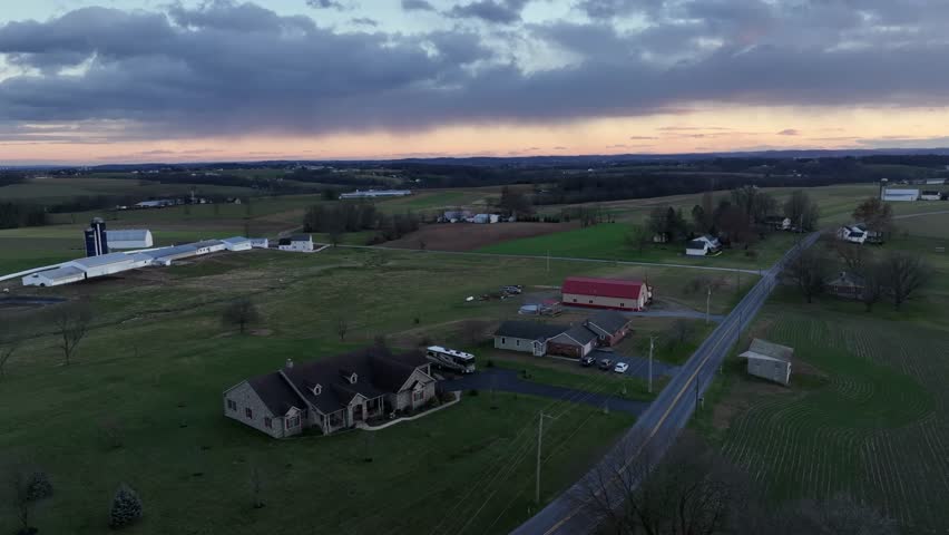 Aerial view of rural American farmland at dusk as open fields, scattered homes and red barn sit under fading sky. Calm countryside atmosphere across quiet United States landscape.