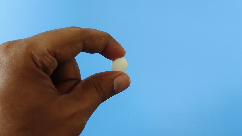 Closeup of asian or indian person hand holding round white medicine pill with finger isolated on blue background and copy space. Painkiller, antibiotic and antidepressant drug bad side effect concept.