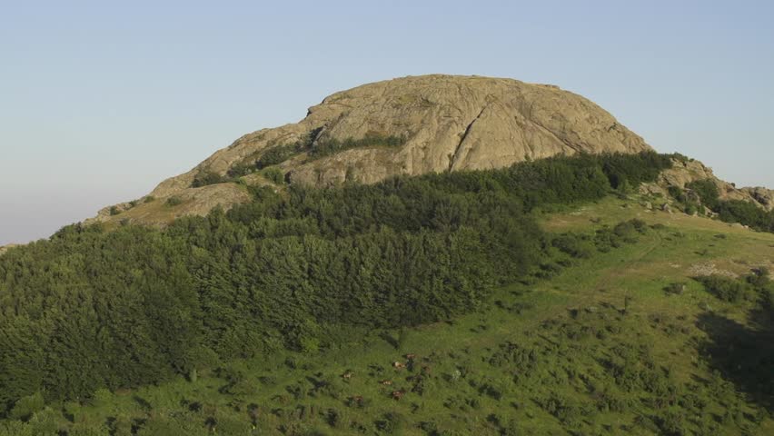 A large hill stands tall in the landscape, covered in grass and surrounded by green trees. Kutelka Reserve in Sinite Kamani Nature Park. Stara planina above Sliven town. Balkan Mountains. Bulgaria.