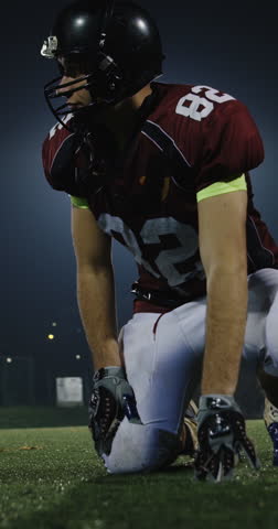 American football player in helmet and maroon jersey kneeling on turf at night, carefully placing the ball for a play under bright stadium lights, focused game preparation