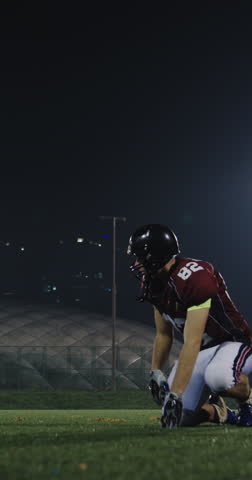 American football player in helmet and maroon jersey kneeling on turf at night, carefully placing the ball for a play under bright stadium lights, focused game preparation
