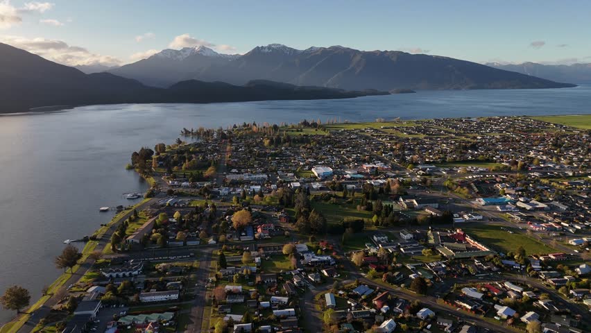 Aerila view of Te Anau Town at sunset with snowy mountains in distance. Sunny day in fall season. Colored trees and tranquil river. Fiordland Region, New Zealand. Wide shot.