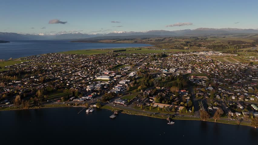 Scenic 4K aerial drone footage of Te Anau town bordering Lake Te Anau in the Fiordland region. Panoramic sunset view of the settlement, water, and distant mountains on South Island