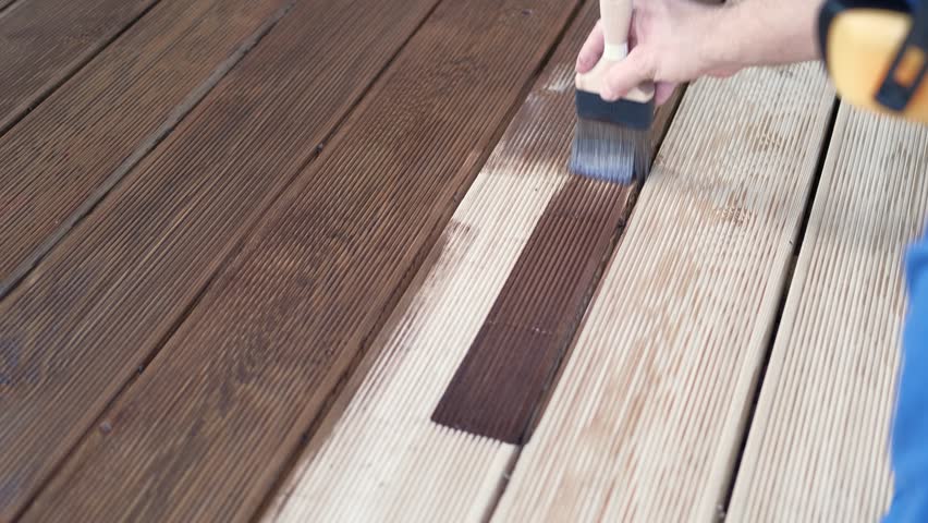 Brown oil for protection of wood by staining. Painting a larch for protecting and pests. Woodworker applying finish to terrace deck board. Repairman varnishing with brush wooden plank floor surface.