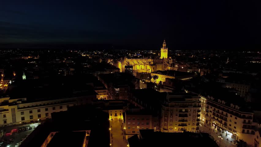 Night aerial shot of Seville city centre with Seville Cathedral and the Giralda bell tower brightly illuminated above the surrounding historic rooftops, Andalusia, Spain