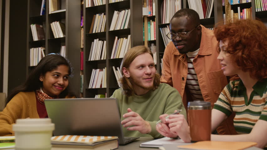 Multi ethnic group of smiling college students discussing homework assignment browsing information on laptop while studying together in library