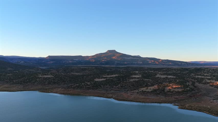 A wide-angle view of the high desert landscape featuring Abiquiu Lake and Cerro Pedernal in northern New Mexico. The terrain is bathed in warm light, characteristic of Georgia O