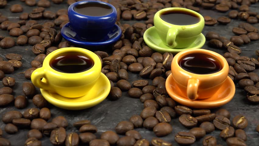 Small colorful cups with hot black coffee, scattered coffee beans are on a black wooden board, side view, rotation