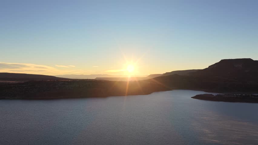 Slow aerial push towards the sun, highlighting the dramatic golden hour light and lens flare. The shot captures the landscape surrounding Abiquiu Lake in New Mexico.