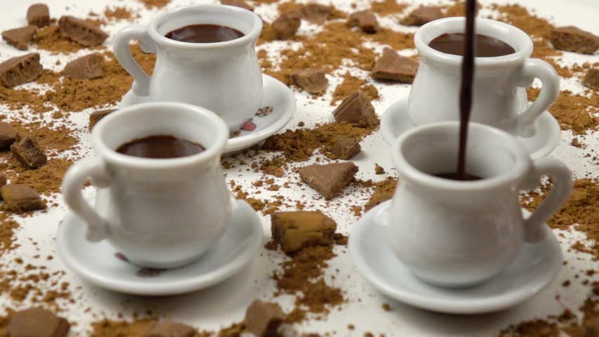 Hot chocolate is pouring in a white cup. White cups with a saucer, pieces of milk chocolate, cacao powder are on a white background, side view, slow motion