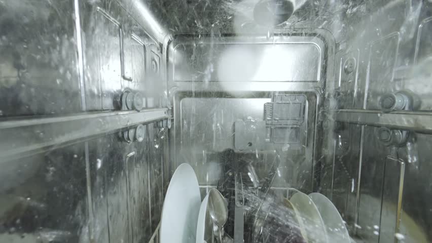 Inside view of a running dishwasher with water spraying over dishes and utensils, creating a dynamic underwater-like perspective from a camera mounted inside the machine