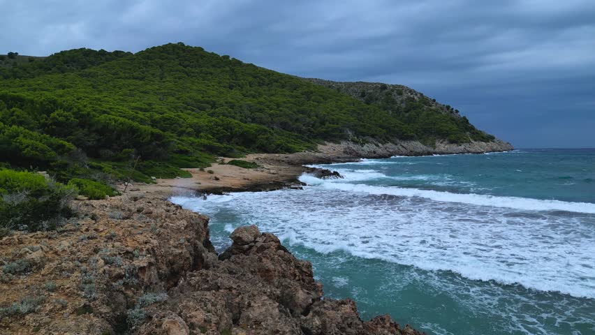 Dramatic high-angle drone shot capturing Cala Agulla beach and its rugged coves in Majorca. Heavy waves under a dark, cloudy sky, highlighting the powerful natural landscape.