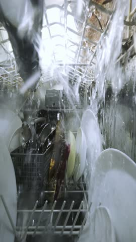 Slow-motion inside view of a running dishwasher with water splashing over plates and utensils, captured from a unique interior camera perspective during the wash cycle