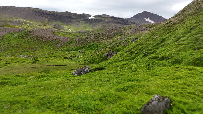 Aerial view of rugged mountains and cliffs in Fjallabyggð, showing rocky slopes, patches of bright green moss, and streaks of lingering snow across a dramatic Icelandic highland landscape