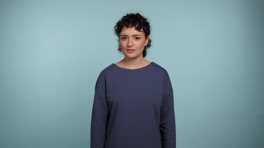 Portrait beautiful smiling curly woman spreads hands says what looks camera. Positive female wearing blue sweater on isolated light blue background with copy space. People emotions lifestyle concept
