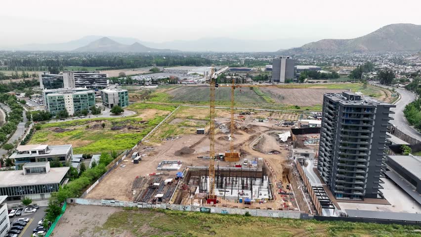 Orbiting drone aerial of construction site with yellow cranes, foundation work, high-rise towers, and mountains valley backdrop panorama