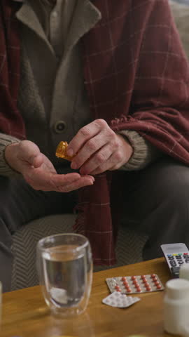 Vertical shot of aged Caucasian man with cold taking pills while sitting on sofa in living room