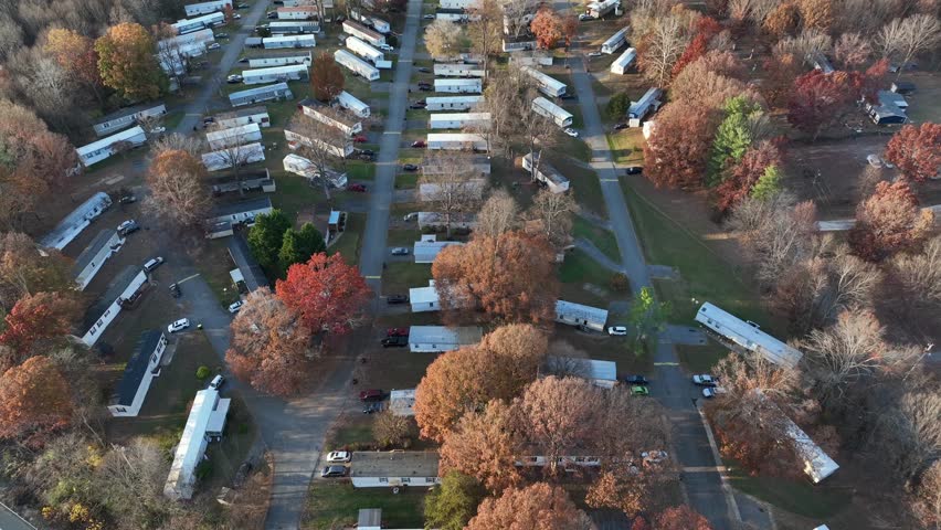 Aeria flyover large American mobile trailer home park set among autumn trees as long rows of white units line narrow roads and quiet community. Wooded landscape in soft daylight in american. top down