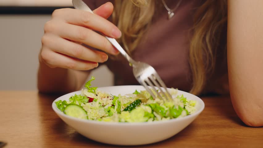 unhappy woman sitting at kitchen table preparing healthy vegetable salad bowl for diet dinner. Gigl mixing salad with fork on plate, bad appetite, sad expression refuses to eat greens healthy eating