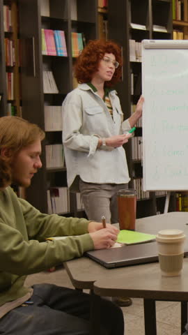 Vertical shot of young woman introducing Spanish phrases to group of adult students gesturing toward whiteboard during beginner level session in library