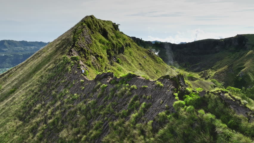 Man running rugged, green covered hills ridge crater rim of Mount Batur volcano, caldera and steaming vents in Bali, Indonesia. Active sports, travel, hiking outdoor. Aerial drone flight panorama