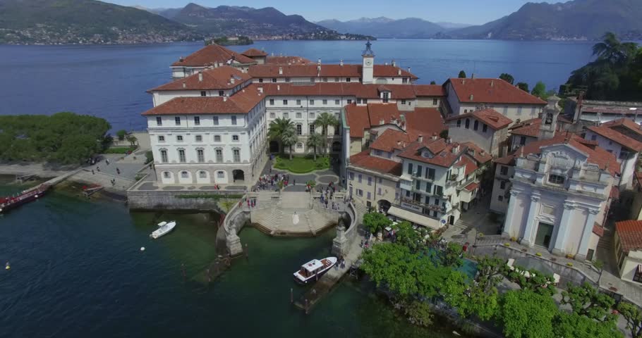Low-angle drone shot of the magnificent Palazzo Borromeo facade on Isola Bella, Lake Maggiore, Italy. The view includes the adjacent historic buildings, the port area with docked tourist boats