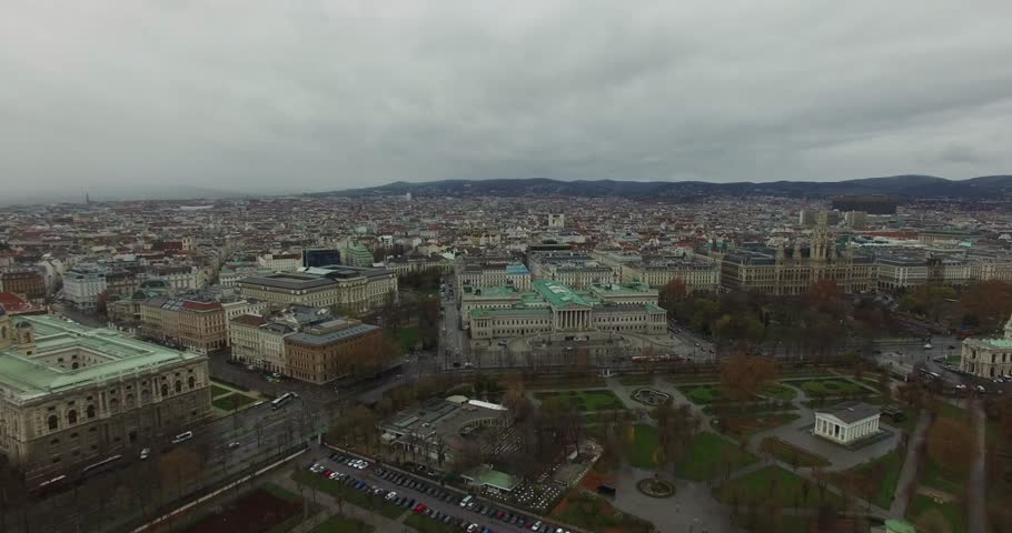 Wide aerial panorama of the historic center of Vienna, Austria, showing a large urban park Volksgarten and massive, neoclassical government buildings Parliament under an overcast, winter sky.