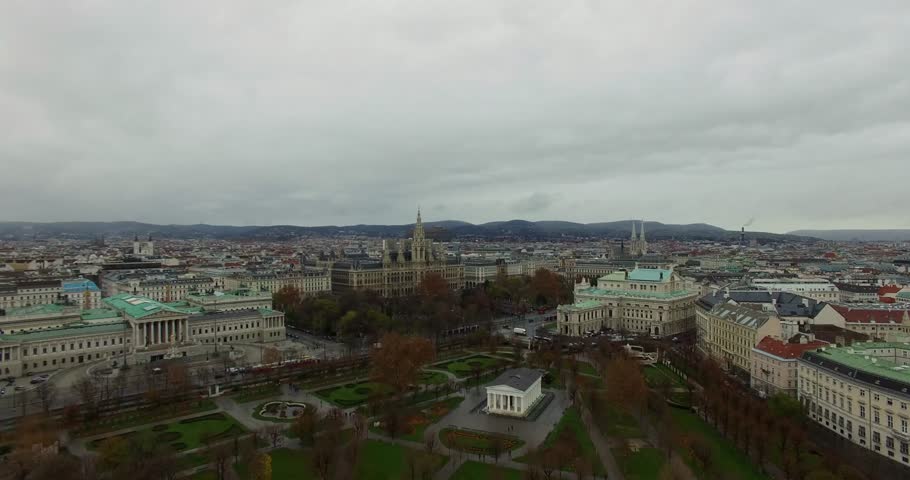 Wide aerial panorama of the central European city of Vienna, Austria, showcasing expansive historic classical architecture, surrounding urban areas, and a large park or green space under an overcast s