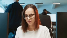 Close camera view of beautiful Caucasian woman facing camera. Female employee giving soft smile. Sitting at desk in bright workspace. In blurred background colleagues focusing on tasks. - Powered by Shutterstock - Get 15% off with code: PIKWIZARD15