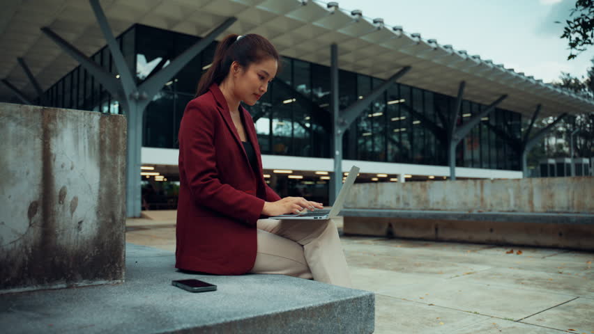 Asian business woman wearing a red suit jacket and using a laptop while sitting on a concrete bench in front of a modern building