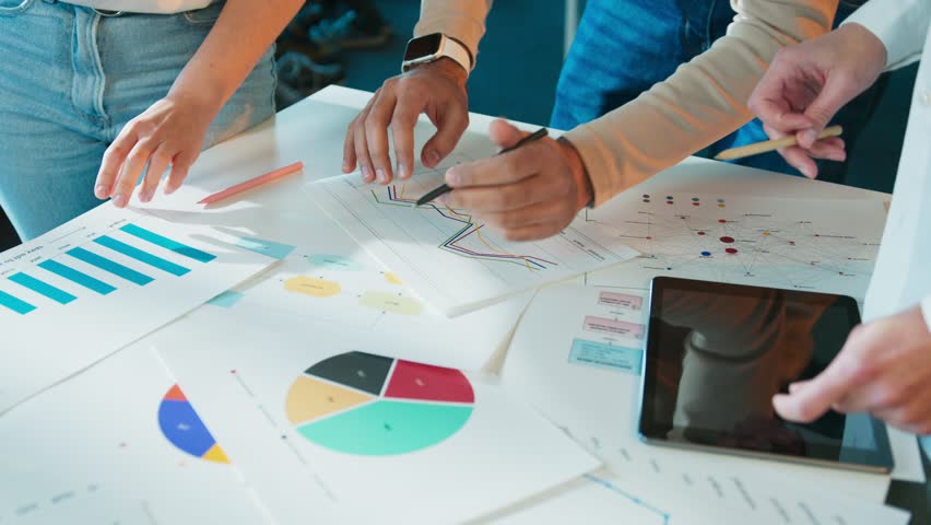 Hands of colleagues leaning over table. Team reviewing printed charts and diagrams. Markers and tablet on surface. Group analyzing workflow steps in office room. Data sheets guiding discussion.