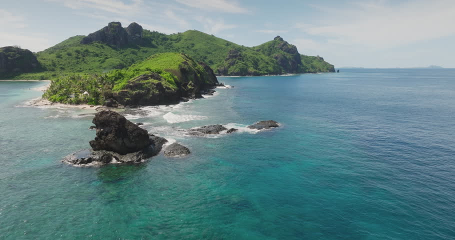 Fiji rocky shoreline with white sand island, turquoise surf breaking over coastal rocks, rugged green mountains under clear blue sky tropical paradise. Beautiful nature landscape. Aerial drone flight