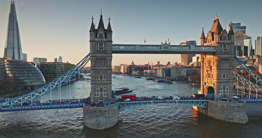 Tower Bridge Victorian Gothic towers and bustling cars traffic cross the River Thames at golden hour sunset, London skyline glowing in warm evening light. Travel destination. Aerial panorama footage