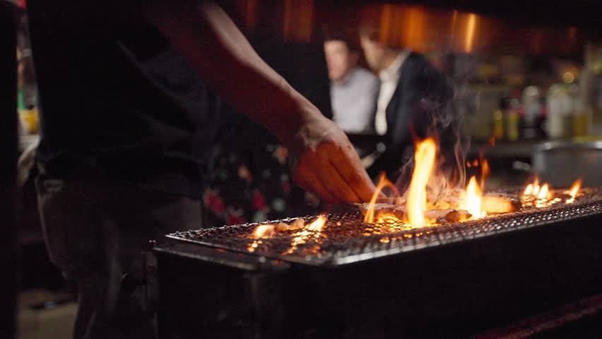 Slow motion of a hand flipping grilled food over open flames at a night yatai in Fukuoka, Japan. The fire flares up while the food is turned during cooking.