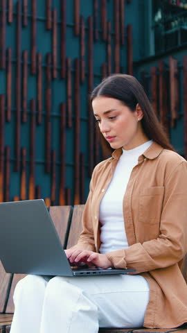 Young business woman remotely on her computer while sitting on bench during lunch break about modern office building, outdoors