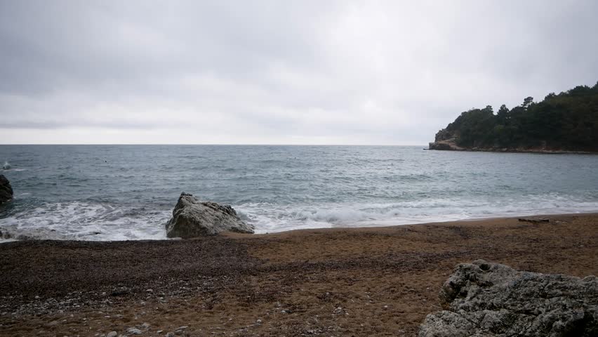 Wide panoramic view of a tranquil seaside landscape under a cloudy, overcast sky. Gentle waves roll onto a sandy shore, with rocky formations and a forested headland visible in the distance. The muted