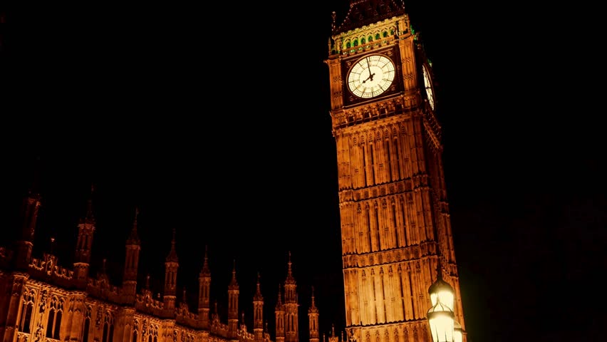 Magnificent clock tower stands tall at night, warmly illuminated against dark sky. Surrounding historic buildings frame this iconic landmark in London, creating a stunning visual display.