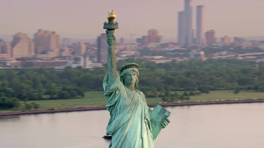Statue of Liberty rises majestically above New York Harbor as sunlight casts warm tones across the city. High view captures bustling surroundings and iconic skyline.