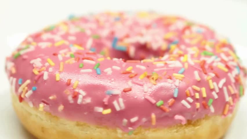 Close up on donut covered in pink frosting and rainbow sprinkles. Sweet pastry rotates slightly against White backdrop. Perfect snack concept. food background