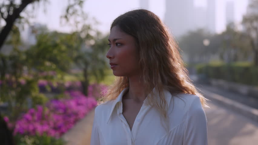 Portrait of young Asian woman in white blouse turns head to camera near purple flowers in garden. Happy female smiles expressing positive vibes in blooming park horizontal shot